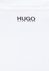Tricou alb cu o textură ribbată, având un logo imprimat negru "HUGO HUGO BOSS" aproape de guler. Design simplu, fără modele suplimentare.