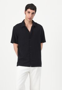 Camicia nera a maniche corte con texture a righe verticali. Presenta un colletto e una vestibilità leggermente rilassata. Abbinata a pantaloni bianchi.