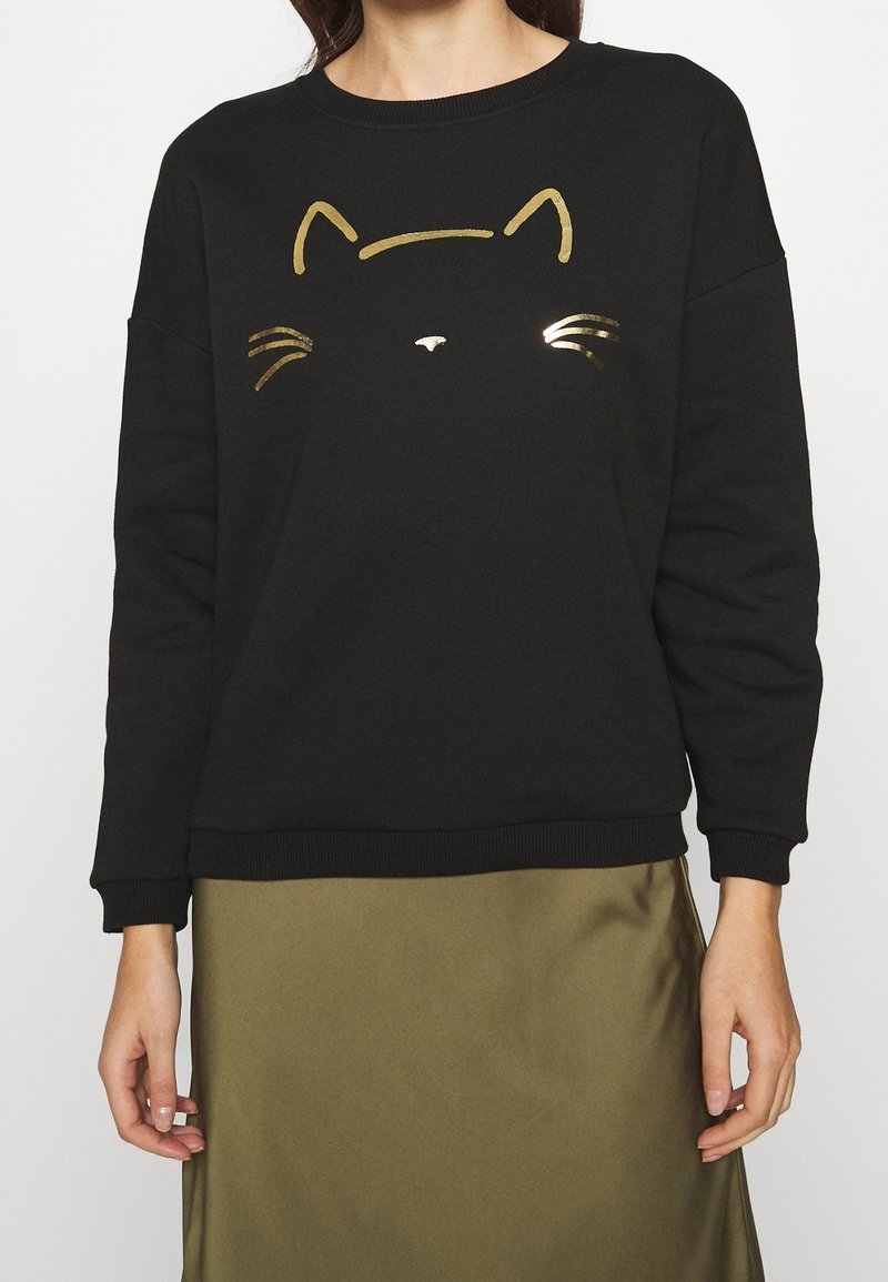 Femme portant un pull à manches longues noir avec un contour doré de visage de chat et une jupe vert olive, montrée du cou jusqu'à mi-cuisse.