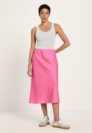 Frau steht und trägt ein ärmelloses graues Oberteil, einen leuchtend pinken Midirock und weiße Turnschuhe vor einem schlichten hellen Hintergrund.