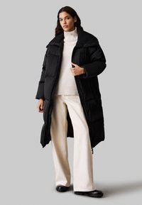 Abrigo acolchado negro con un cuello alto y corte oversize, diseñado sobre un jersey de cuello alto crema y pantalones anchos crema, complementado con calzado negro.