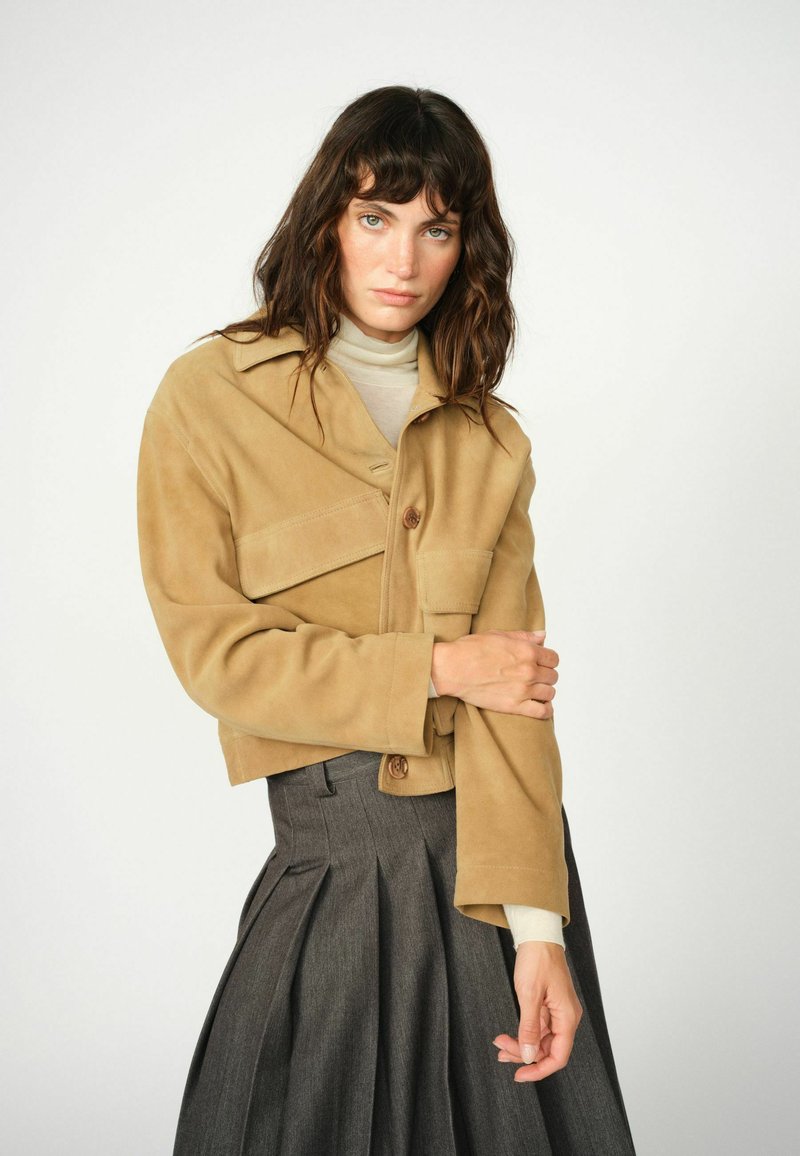Tan suede cropped jasje met een kraag, enkele knoopsluiting en een borstzak. Ggedragen over een lichte trui en een grijze geplisseerde rok.