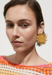 Gouden oorknoppen in de vorm van zonnebloemen met textuurdetails; model draagt een kleurrijke, opengehaakte top met roze, oranje en gele tinten.