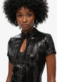 Haut noir paillettes avec un col haut, des manches courtes, une découpe en œil de serrure et des liens décoratifs avec des accents en perles. Texture lisse et réfléchissante.