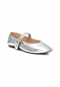 Zapato plano metálico plateado con punta afilada y una correa decorativa con hebilla y agujeros, superficie texturizada y tacón mínimo.