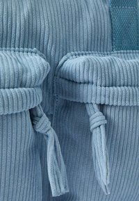 Hellblaues Cordtasche mit zwei vorderen Taschen mit geknoteten Verschlüssen und strukturiertem senkrechtem Rippmuster. Akzentuiert mit einem flachen Griff.