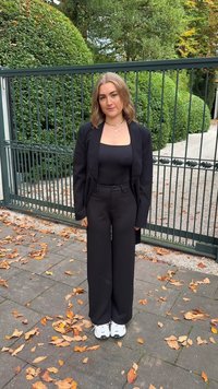 Μαύρο προεξέχον σακάκι πάνω από μαύρο τοπ, συνδυασμένο με μαύρα παντελόνια με φαρδιά πόδια. Λευκά sneakers ολοκληρώνουν το σύνολο. Φύλλα στο έδαφος.