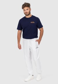 Marineblaues T-Shirt mit orangefarbenem Logo, weiße Hose mit elastischem Bund und Seitentaschen sowie weiße Turnschuhe. Lässige Sportbekleidung.