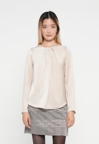 Halenka s dlouhým rukávem v měkké béžové tkanině s detailním skládáním v přední části, spárovaná se šedou sukní s houndstooth vzorem. Hladká textura, kulatý výstřih.