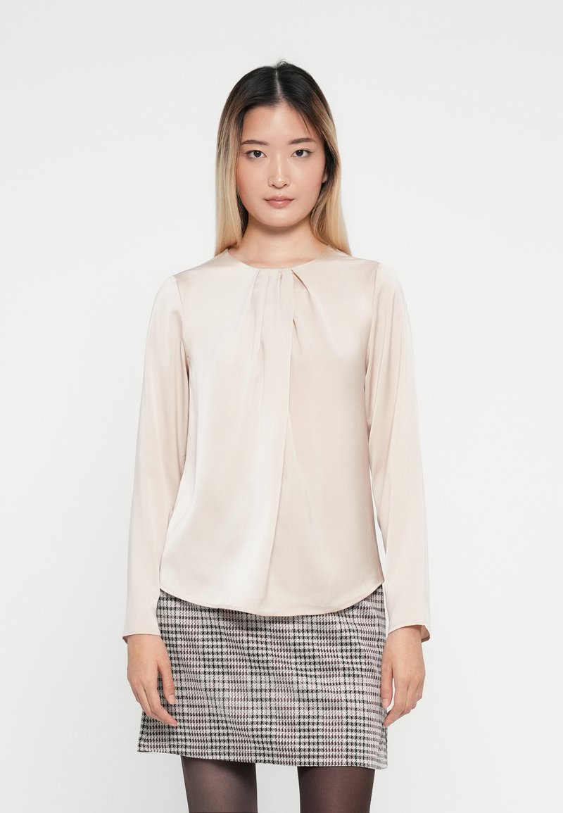 Halenka s dlouhým rukávem v měkké béžové tkanině s detailním skládáním v přední části, spárovaná se šedou sukní s houndstooth vzorem. Hladká textura, kulatý výstřih.