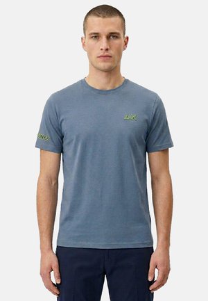 Uomo con t-shirt blu a maniche corte con logo verde "ΔX" sul petto e testo "SUN 68" sulla manica sinistra, in piedi contro uno sfondo semplice.