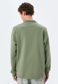 Grünes Langarmhemd mit geripptem Kragen, aus weichem Stoff, mit lockerem Schnitt und minimalistischem Design, von hinten gezeigt.