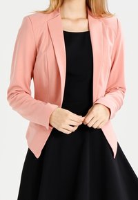 Blazer rose pâle en tissu lisse avec une coupe ajustée, col à revers crantés et fermeture par un bouton, assorti à une robe noire.