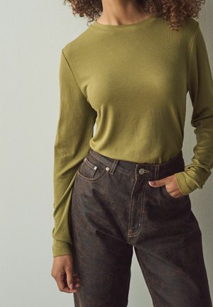 Femme portant un haut côtelé à manches longues vert olive rentré dans un pantalon texturé foncé taille haute, main dans la poche, debout contre un mur clair.