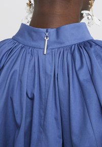 Gros plan sur une fermeture éclair au dos d'une robe plissée bleue portée par une personne à la peau foncée avec des boucles d'oreilles florales blanches.