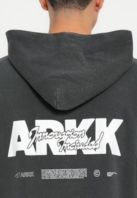 Hallhallane kapuutsiga, millel on suur valge logo "ARKK" ja fraas "Innovatsioon kaasatud" seljaosas, lõ relaxed fit ja tekstuuriga kangas.