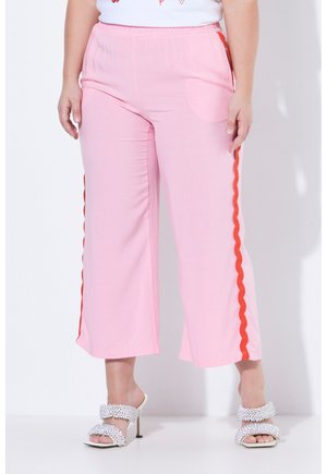 Personne grande taille portant un pantalon rose à jambes larges avec des rayures ondulées rouges sur les côtés et des sandales blanches à talons hauts avec des ornements.