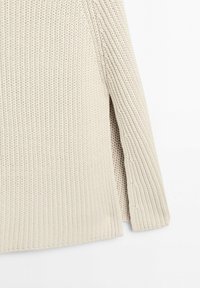 Beżowy, gruby sweter z dzianiny o wyraźnej prążkowanej fakturze i bocznym rozcięciem na dolnym brzegu na białym tle.