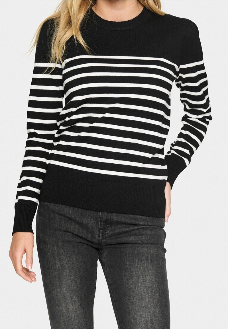 Femme portant un pull rayé horizontal noir et blanc à manches longues et un jean gris foncé, debout avec une main dans la poche.