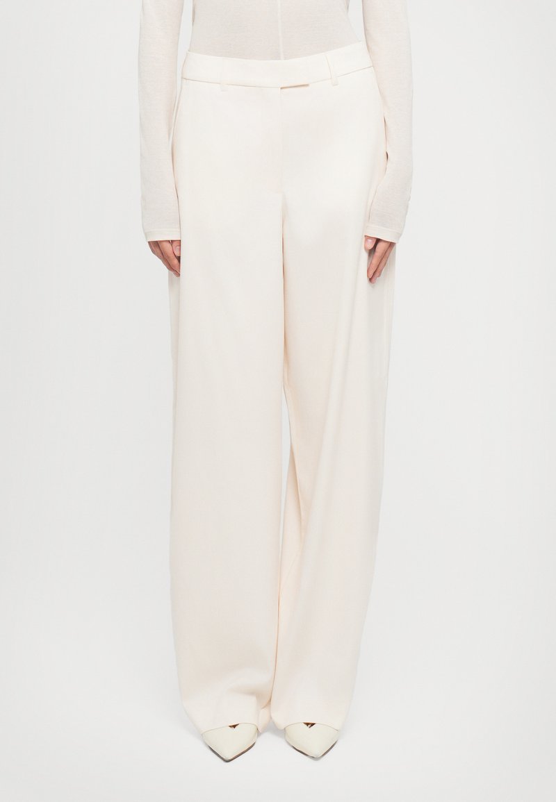 Donna che indossa ampi pantaloni beige a vita alta con passanti per cintura, abbinati a un top beige a maniche lunghe e scarpe puntate color crema.