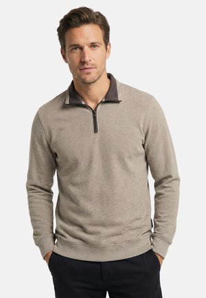 Mand iført beige quarter-zip sweater med mørkræv og sorte bukser, stående med hænderne i lommerne mod hvid baggrund.