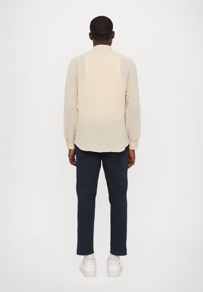 Camicia a maniche lunghe beige chiaro in tessuto texturizzato, con orlo curvato, abbinata a pantaloni blu navy e scarpe da ginnastica bianche, vista da dietro.