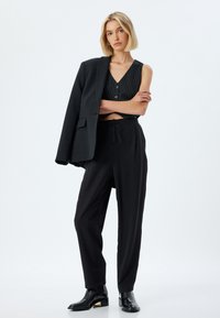 Blazer noir ajusté posé sur un top sans manches noir à rayures et un pantalon noir ample. Bottines noires brillantes avec des accents métalliques.