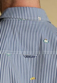 Prúžkovaná košeľa v modro-bielej farbe z bavlny s malými vyšívanými ikonami (miska, syr) na zadnej strane, s klasickým golierom a jemným logom.