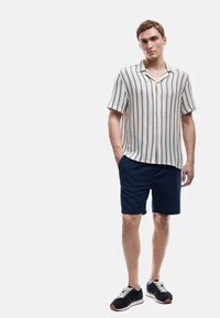 Camisa de manga corta color crema con rayas azules, combinada con pantalones cortos azul marino. El modelo lleva zapatillas negras con cordones blancos y suela clara.