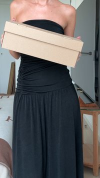 Scatola di cartone per scarpe tenuta con entrambe le mani, forma rettangolare, colore marrone chiaro, texture liscia, con un design semplice del coperchio. Un abbigliamento nero pende sotto.