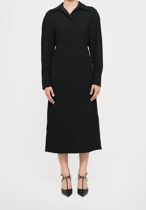 Femme portant une longue robe noire à col avec manches longues et des chaussures à talons noirs avec brides à la cheville sur fond blanc.