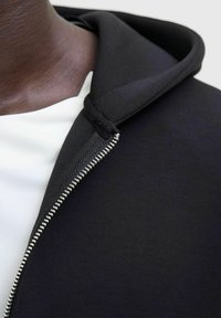 Sort hætte sweatshirt med lynlås fortil, som har et struktureret stof og en glat indvendig side. Lynlåsen er metallisk, hvilket står i kontrast til det mørke materiale.