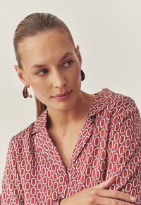 Cămașă roșie cu model și legături geometrice albastre; cercei maro lucioși cu accente aurii; material neted; detalii ale gulerului și nasturilor vizibile.