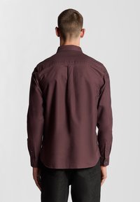 Camicia a maniche lunghe color bordeaux con orlo dritto e polsini abbottonati. Tessuto liscio con dettagli di plissè sul retro. I polsini regolabili presentano bottoni neri.