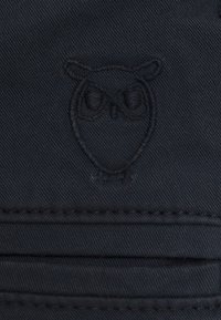 Svart tyg med ett strukturerat uggle-emblem, sydd med fina detaljer. Materialet verkar hållbart med synliga sömmar.