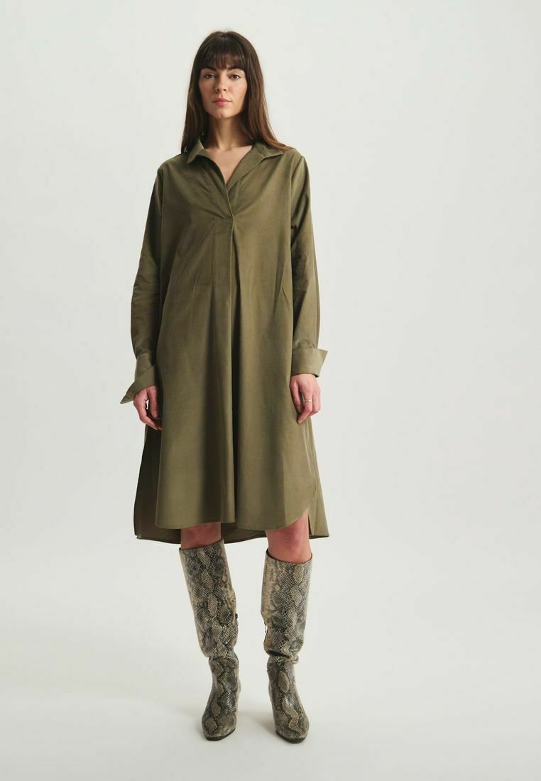 Robe chemise vert olive avec un col, des manches longues et un devant plissé. Associée à des bottes en fausse peau de serpent à motif, montant jusqu'aux genoux.