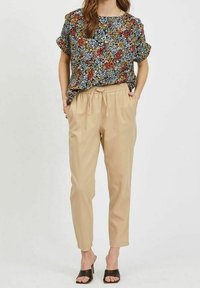 Nainen, jolla on lyhythihainen kukkapaidat, beige narupöksyt ja mustat avokkaat, seisoo kädet taskuissa.