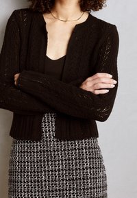 Svart stickad cardigan med ett strukturerat mönster, ribbade ärmslut och öppen fram, i kombination med en svart topp och en mönstrad vävd kjol.