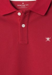 Rotes Polohemd aus strukturiertem Baumwollmaterial, mit klassischem Kragen, drei Knöpfen und einem weißen gestickten Logo auf der linken Seite.