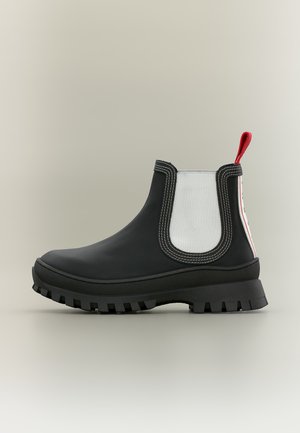 Bottine Chelsea noire avec semelle épaisse, panneaux latéraux élastiques blancs et languette rouge sur fond gris clair.