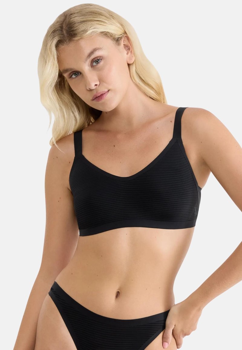 Γυναίκα με ξανθά μαλλιά που φοράει μαύρο ριμπ ανώτερο μέρος και ταιριαστό σλιπ, σε απλό φωτεινό φόντο.