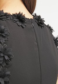 Robe noire à l’arrière avec fermeture éclair et applications florales en tissu le long du décolleté et des épaules sur une personne à la peau claire et aux cheveux bruns.