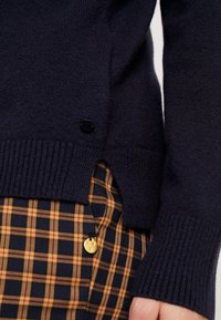 Marineblauwe gebreide trui met ribbelrand en zijsplitten, voorzien van een zwart knoopdetail. Gecombineerd met mosterd- en marineblauwe geruite broek.