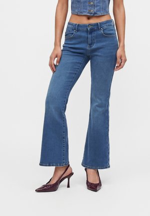 Kvinde iført blå højtsiddende flare jeans, cropped denim vest og spidse mørkerøde slingback hæle, stående mod en ensfarvet baggrund.