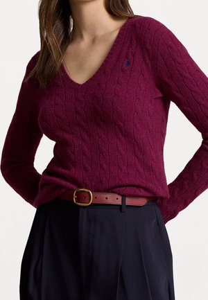 Femme portant un pull col en V prune foncé à torsades, rentré dans un pantalon noir taille haute avec une ceinture bordeaux à boucle dorée.