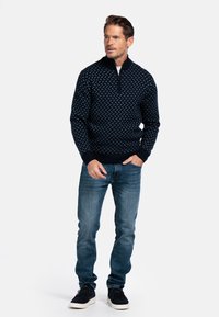 Navy zip-up trui met een geometrisch lichtblauw patroon, textuurgebreide stof, geribbelde manchetten, gecombineerd met lichtblauwe jeans en zwarte sneakers.