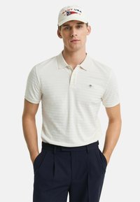Polo crème rayé avec un col, associé à un pantalon bleu marine. Une casquette légère avec le logo "GANT USA" vient compléter la tenue.