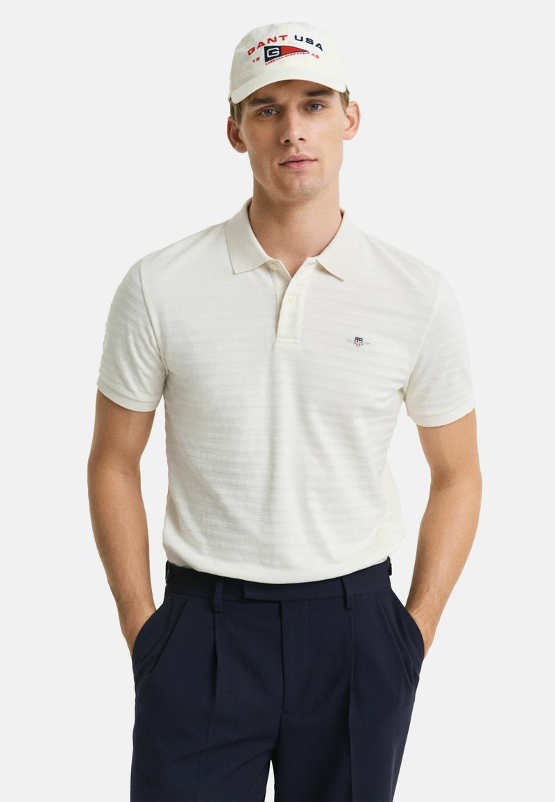Polo crème rayé avec un col, associé à un pantalon bleu marine. Une casquette légère avec le logo "GANT USA" vient compléter la tenue.