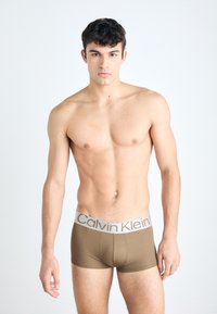 Smeđe pamučne bokser gaće s sivim Calvin Klein pojasom. Model stoji licem prema naprijed s neutralnom pozadinom.