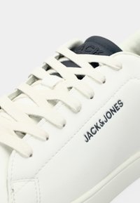 Baskets blanches avec lacets blancs et logo noir "JACK & JONES" sur le côté, avec une languette noire.